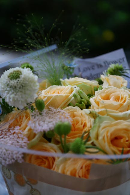 A close-up of a floral gift arrangement featuring soft peach-colored roses with lush petals, complemented by delicate white scabiosa flowers and small green buds. The bouquet is wrapped in beige paper