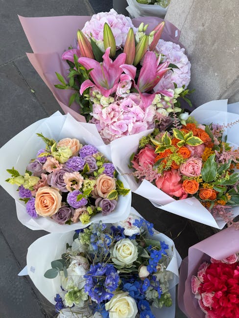 A collection of five beautifully arranged flower bouquets, each wrapped in pastel pink, white, and purple tissue paper, displayed on a grey surface. The top bouquet features a mix of pink lilies with 