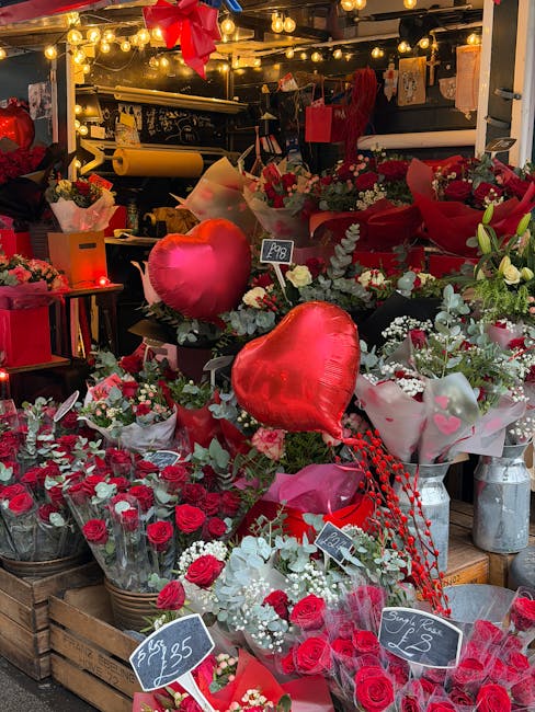 A vibrant display of floral arrangements outside a florist shop for Valentine's Day, featuring red roses, white fillers, and greenery. Large red heart-shaped balloons are integrated into the display, 