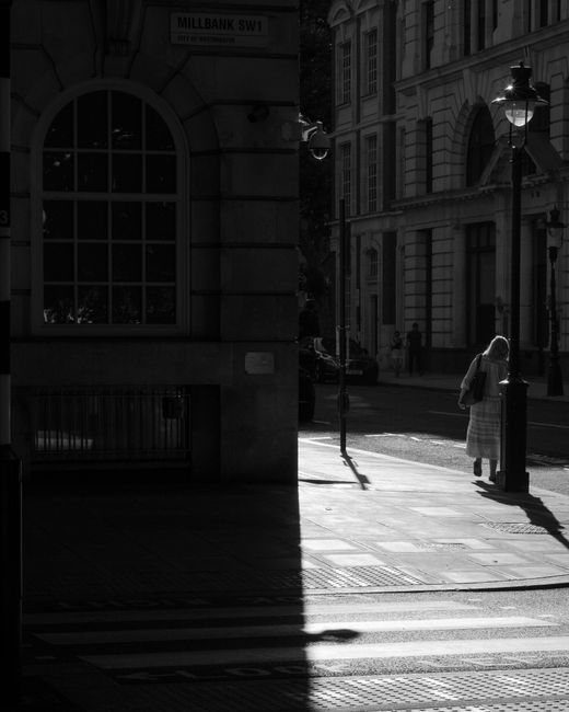 The image features a woman standing on a city sidewalk near Westminster Bridge, illuminated by sunlight with shadows cast across the pavement. She is dressed in a long, checked dress and carries a bag