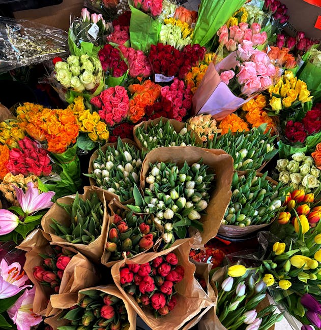 A vibrant display of fresh floral bouquets arranged tightly together, featuring a variety of flowers including pink, yellow, orange, red, and white tulips, roses, and other blossoms. The bouquets are 