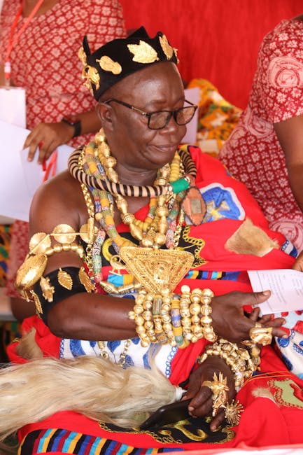 A woman dressed in traditional Maasai attire with vibrant red, blue, and black fabric, wearing elaborate gold jewelry including necklaces, bracelets, and a beaded headband, seated indoors against a re