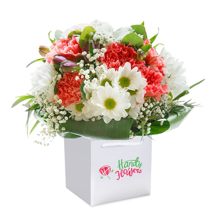 Brimming with artistry and freshness, this thoughtfully crafted bouquet stands tall within a sleek, square white gift bag-its silhouette sharply defined by the studio-white background, reminiscent of Millbank's gallery spaces. The arrangement bursts with a harmony of soft coral and peach carnations whose ruffled petals create a lush, textured core, punctuated by lively, vibrant red blooms for added depth. Dotted between are brilliant white daisies with vivid lime-green centers, their cheerful faces reminiscent of classic springtime in Millbank's riverside parks. Floating among the primary blooms, delicate sprigs of baby's breath add a softening, cloud-like touch-lending an airy delicacy that evokes the gentle breeze from the nearby Thames. On one side, a cluster of purple-tinged buds peeks out, enhancing the bouquet's dimension with subtle, mysterious allure. Rich, glossy green foliage arcs around the base, framing the flowers with natural elegance and structure, offering both support and an appealing visual backdrop. The overall composition forms a generous, rounded mound, brimming with freshness and intent-each stem expertly placed as if curated for an artful display along the streets of Millbank. The pristine white gift bag, detailed with robust ribbon handles, ensures easy carrying to local celebrations, be it an anniversary near Millbank's institutions or a birthday surprise. This bouquet, photographed with expert lighting to capture its pure hues and textures, embodies the refined charm and heartfelt warmth that characterise floral gifts delivered throughout the community.